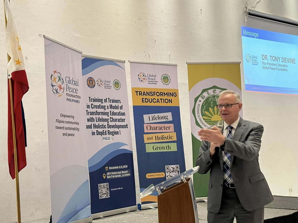A man in a suit speaks at a podium. Behind him are banners for Global Peace Foundation, featuring educational themes like lifelong learning, character, and holistic growth.