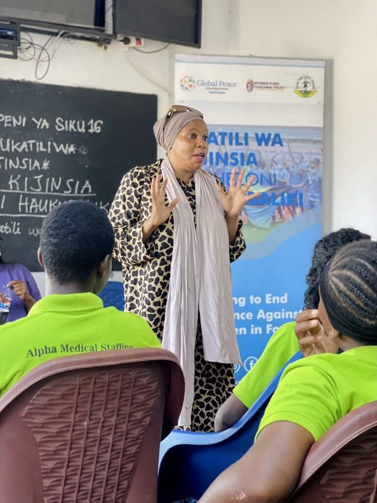 In a classroom adorned with a banner and chalkboard, a woman in a spotted dress addresses attendees in green uniforms, emphasizing the importance of strengthening communities. The event is part of GPF Tanzania's initiative to empower youth through education and collaboration.