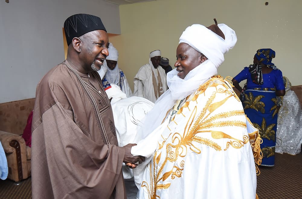 Two men in traditional attire are shaking hands in a room, embodying peace and inclusivity. One wears a dark outfit with a hat, the other a white and gold robe with a turban. Background figures observe this harmonious exchange, reminiscent of Nigeria’s diverse cultural tapestry.