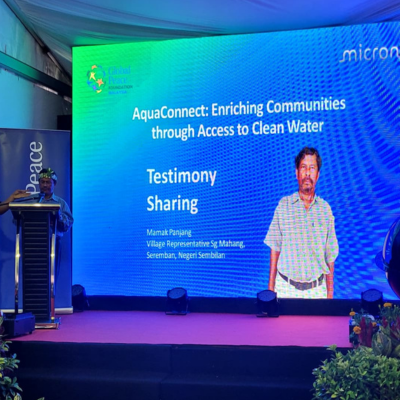 A speaker on stage stands next to a large screen displaying a slide titled "Testimony Sharing" with the topic "AquaConnect: Enriching Communities through Access to Clean Water," highlighting how Micron and Global Peace partner to bring clean water to Orang Asli communities.