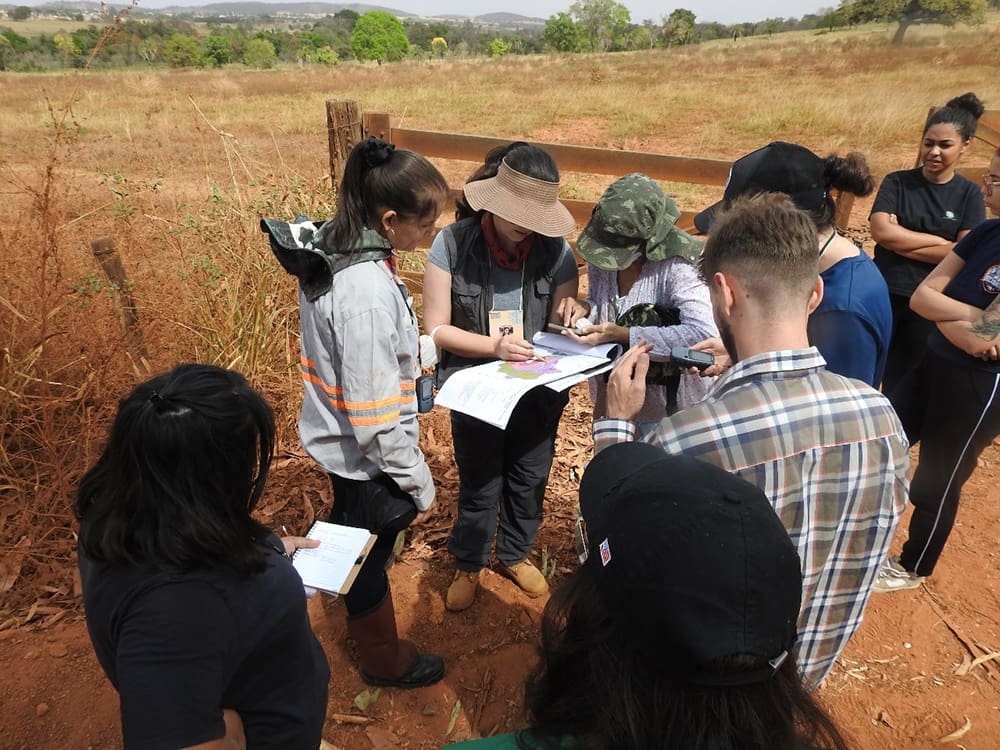 A group of people, embodying the spirit of community empowerment, stands outdoors in a dry field, examining documents and taking notes. Some are wearing hats and casual clothing, totally engaged in their mission.