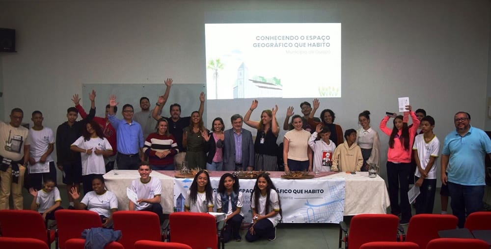 In a dynamic lecture hall, a diverse group eagerly engages, some with hands raised high. Behind them, a screen reads