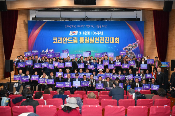 A large group of people hold blue and purple signs in a theater with a blue banner behind them, promoting the spirit of the March 1st Movement. The audience watches from red seats in the foreground, captivated by this moving tribute to unity.