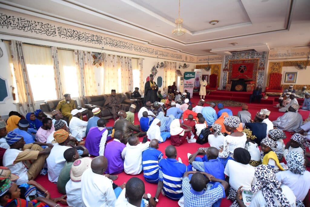 A large gathering of people from Agaie LGA seated on a red carpet in a decorated hall in Niger State, Nigeria, facing a panel of speakers and display banners.