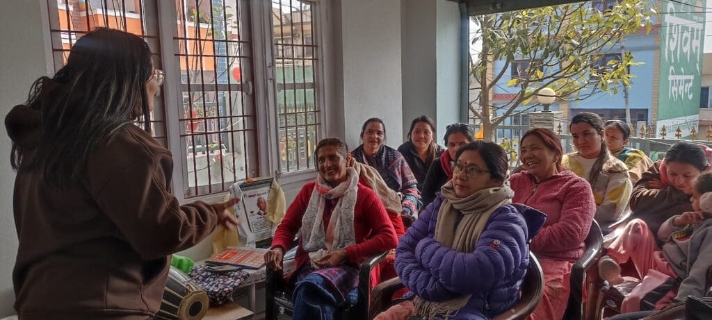 A person stands speaking to a seated group in a sunlit room, their audience warmly dressed and attentive. The conversation centers around women empowerment, sparking engagement and inspiration among all present.