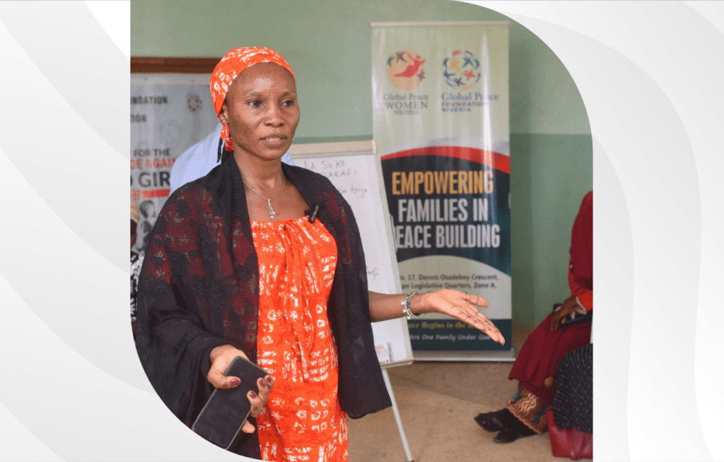 A woman in traditional attire speaks passionately, gesturing with her hand. Behind her, a banner celebrates Women in Peacebuilding, emphasizing their crucial role in empowering families. The scene highlights the spirit of International Women’s Day 2025.