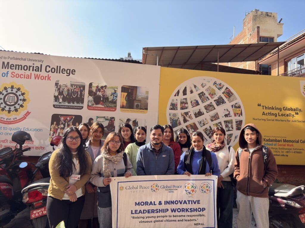 A group of people stands in front of a college building in Nepal, proudly holding a banner for a Moral & Innovative Leadership Workshop. The scene is alive with educational posters and a brick wall backdrop, embodying the spirit of supporting youth leadership.