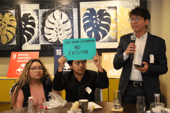 Three people sit at a table in a meeting room adorned with framed art and printed signage. One holds a sign saying, 