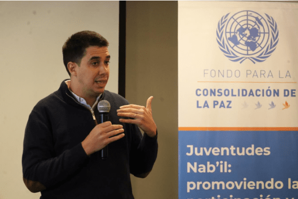 A person speaking into a microphone stands beside a banner with the United Nations emblem and text in Spanish about peace consolidation and youth participation, reflecting initiatives supported by the Global Peace Foundation in Central America.