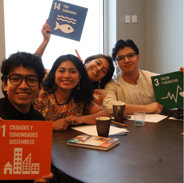 Four people sitting at a table, holding sustainable development goal signs, and smiling at the camera exude positivity and unity. Their collaboration reflects the mission of the United Nations to promote global peace and progress.