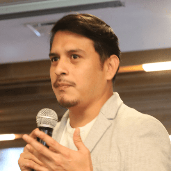 A man holding a microphone and speaking passionately about the Global Peace Foundation, wearing a light gray blazer, stands against a blurred background of wooden panels and lights.
