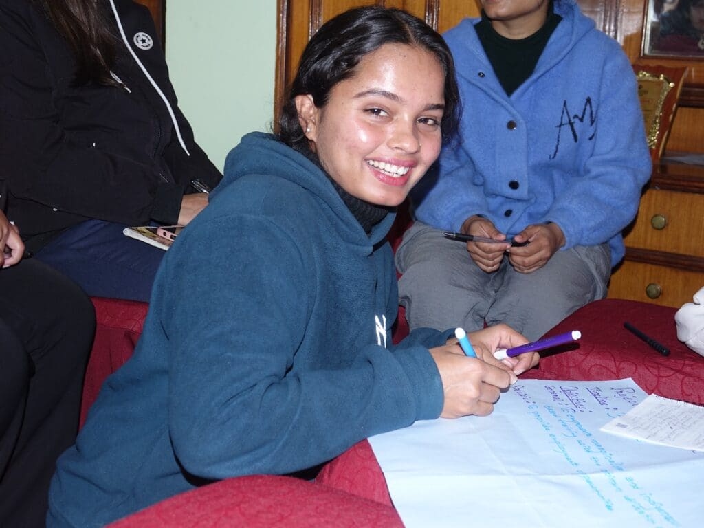 A person in a blue hoodie smiles while holding pens and writing on a large sheet of paper indoors, embodying youth leadership. Nearby, others are seated, engaged in supporting youth initiatives that foster positive change.