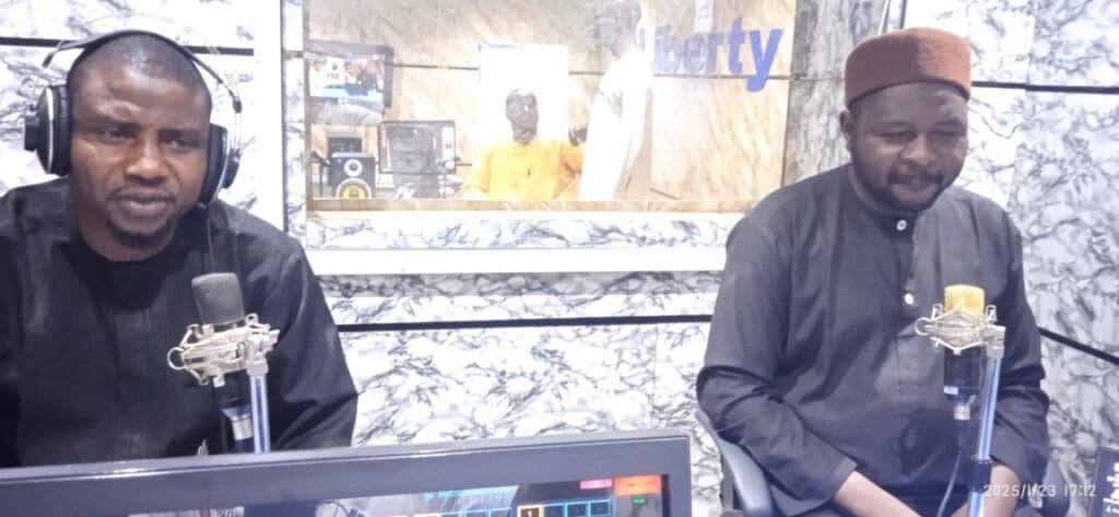 Two men wearing headphones speak into microphones in a radio studio, broadcasting a lively radio program. A person is visible in the background through a window, embodying the sense of unity akin to