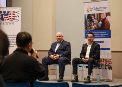 Two men sit on a stage in front of banners during the Korean Dream campus tour, facing an audience member taking a photo. The backdrop displays "Global Peace Foundation" and "USA" with a Korean translation.