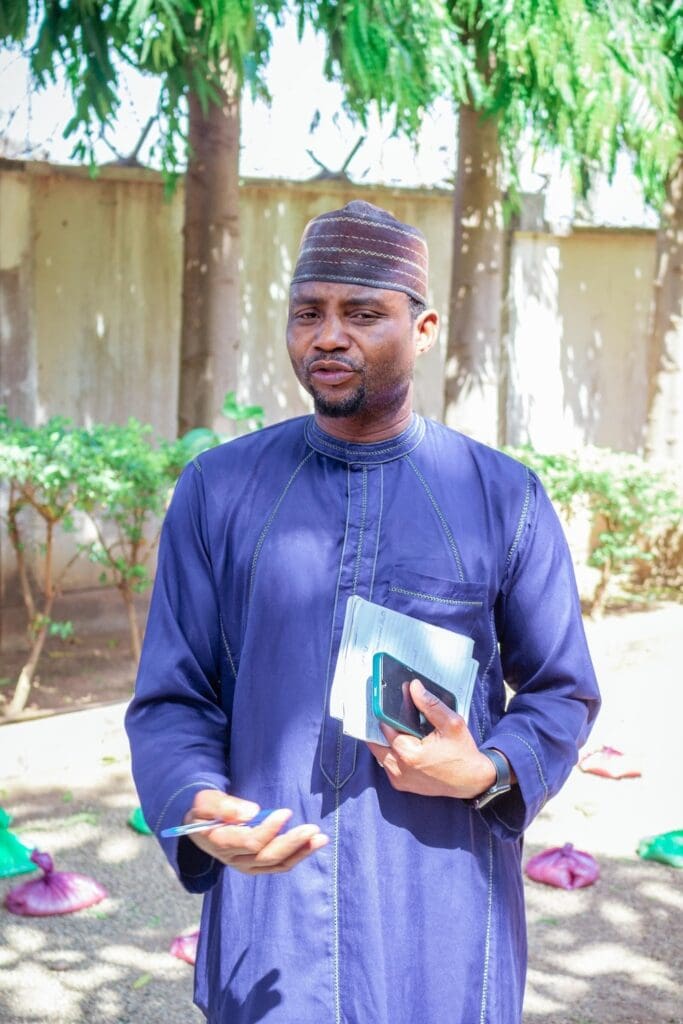 A man in a blue traditional outfit stands outdoors, promoting interfaith harmony as he holds documents and a phone, with green trees and small bags visible in the background.