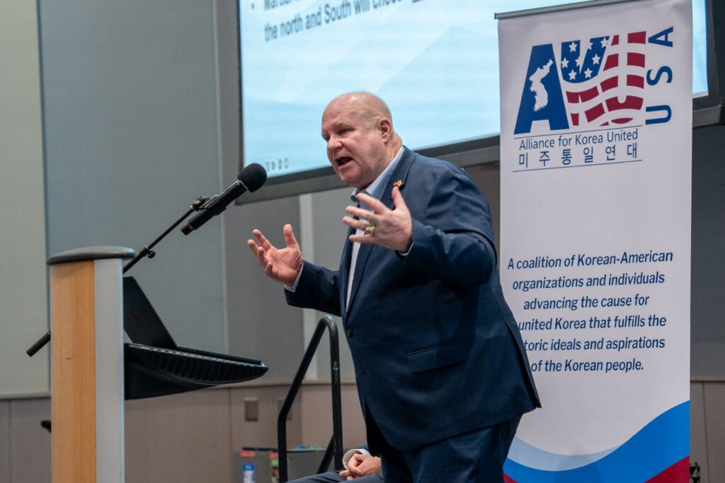 A man speaks at a podium with a microphone as the banner beside him displays the Alliance for Korea United logo, emphasizing the Korean-American cooperation. The event is part of the inspiring Korean Dream campus tour, promoting unity and shared vision.