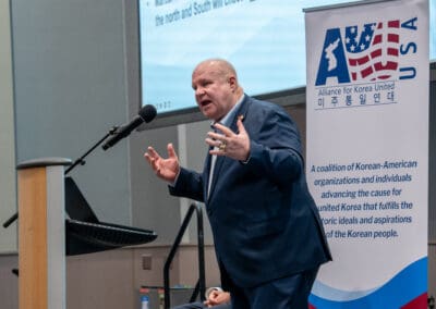 A man speaks at a podium with a microphone as the banner beside him displays the Alliance for Korea United logo, emphasizing the Korean-American cooperation. The event is part of the inspiring Korean Dream campus tour, promoting unity and shared vision.