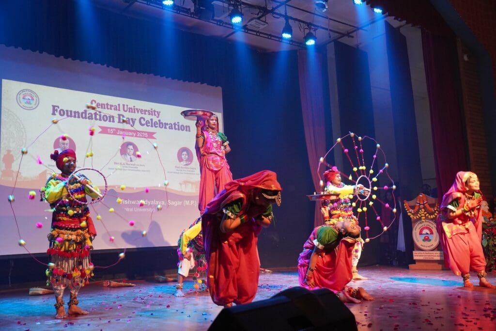 Performers in colorful traditional attire dance on stage at the Central University Foundation Day Celebration, their graceful movements embodying a mission of spreading peace, set against a backdrop displaying event details at Dr. Harisingh Gour Vishwavidyalaya.
