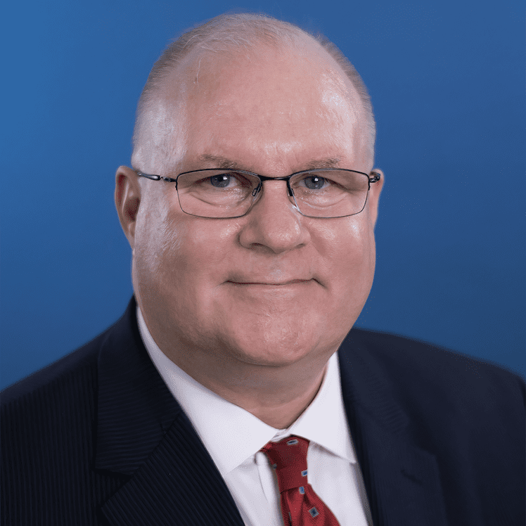 A man in a suit with a red tie is smiling against a blue background. He is wearing glasses and has short, light-colored hair.