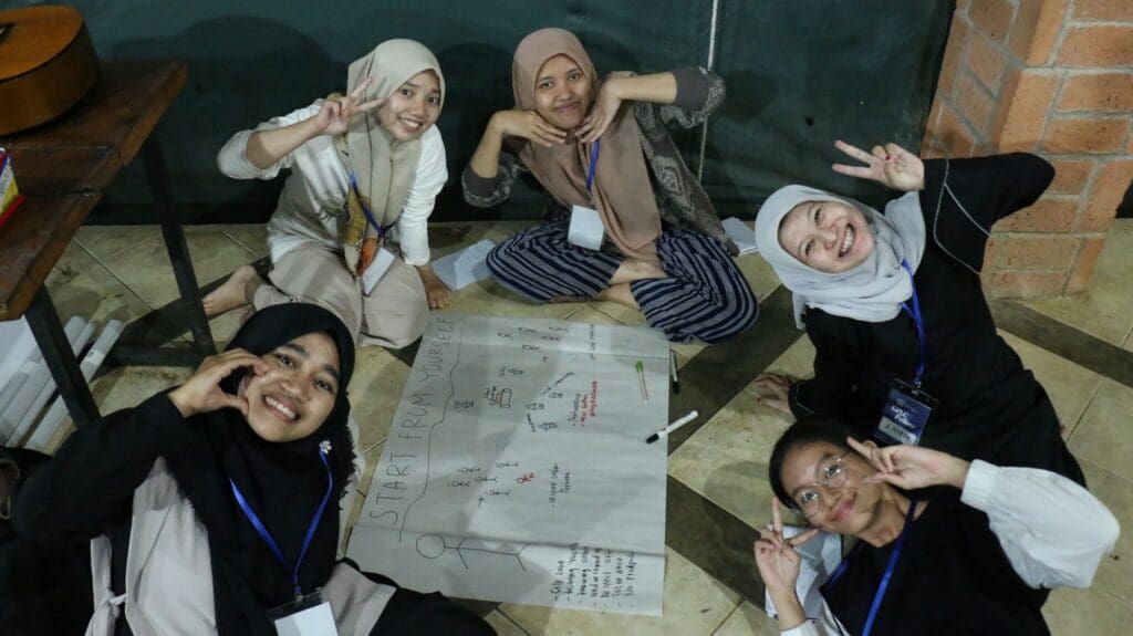 Five young women, aspiring peacebuilders from the Global Peace Leadership Corps Indonesia, sit on the floor around a large paper filled with diagrams and notes, posing and smiling at the camera.