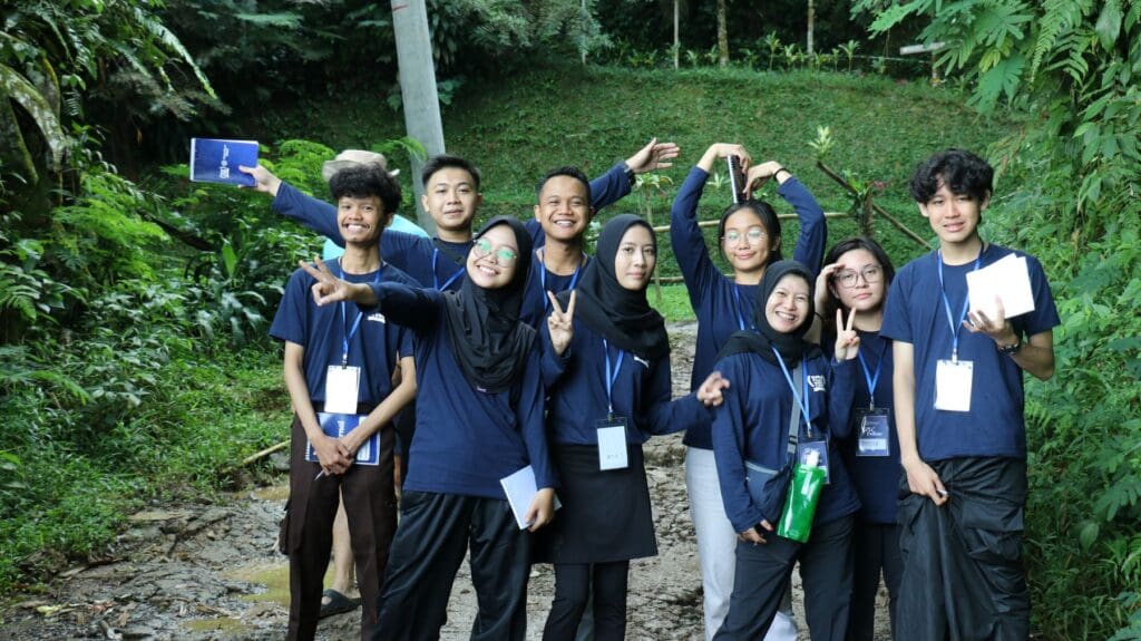 A group of nine young peacebuilders from Global Peace Leadership Corps Indonesia, wearing matching navy shirts and name tags, pose together on a forest path, smiling and making hand signs.