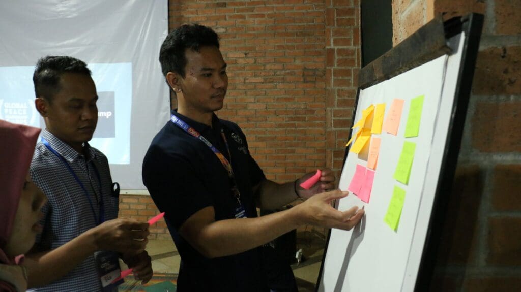Two people stand by a whiteboard, placing colorful sticky notes on it, as part of a Global Peace Leadership Corps Indonesia group activity in an indoor setting with brick walls.