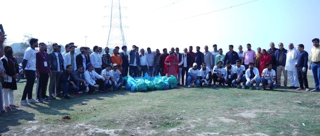 A large group of people stands on grass near trash bags filled with rubbish, participating in the 2025 Yamuna River Clean-Up. A power line tower is visible in the background.