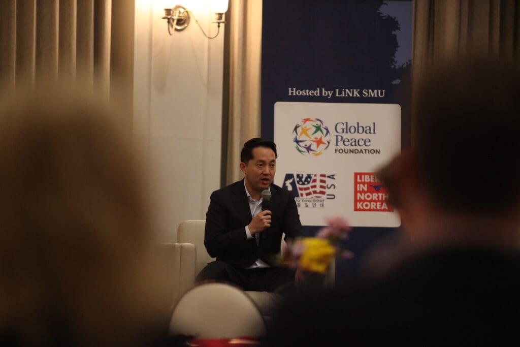 A person speaks into a microphone while seated on a stage, part of the Korean Dream campus tour. A backdrop displays logos for the Global Peace Foundation and other organizations, capturing the attentive audience members visible in the foreground.