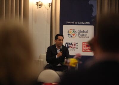 A person speaks into a microphone while seated on a stage, part of the Korean Dream campus tour. A backdrop displays logos for the Global Peace Foundation and other organizations, capturing the attentive audience members visible in the foreground.