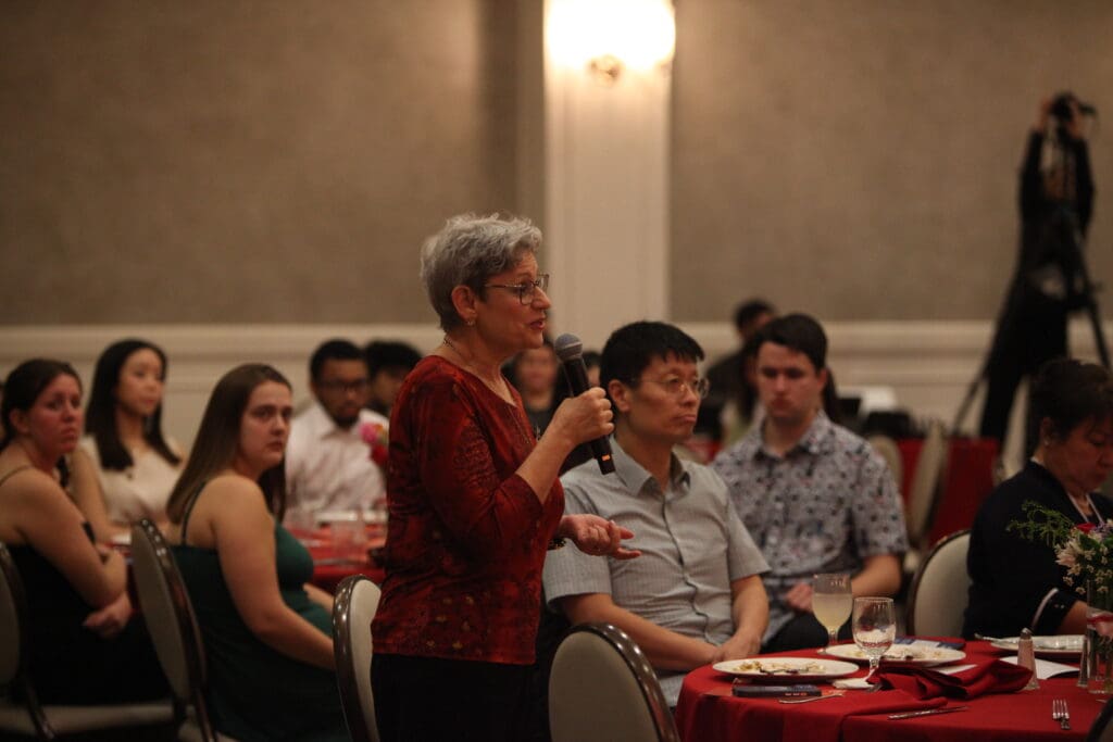 An older woman in a red blouse speaks into a microphone at a banquet, sharing her inspiring journey from the Korean Dream to a campus tour with attentive attendees surrounding her.
