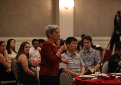 An older woman in a red blouse speaks into a microphone at a banquet, sharing her inspiring journey from the Korean Dream to a campus tour with attentive attendees surrounding her.