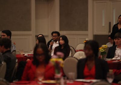A man holding a microphone is seated among attendees at a formal event, complete with red tablecloths and floral centerpieces, possibly narrating his vibrant experiences from the Korean Dream campus tour.