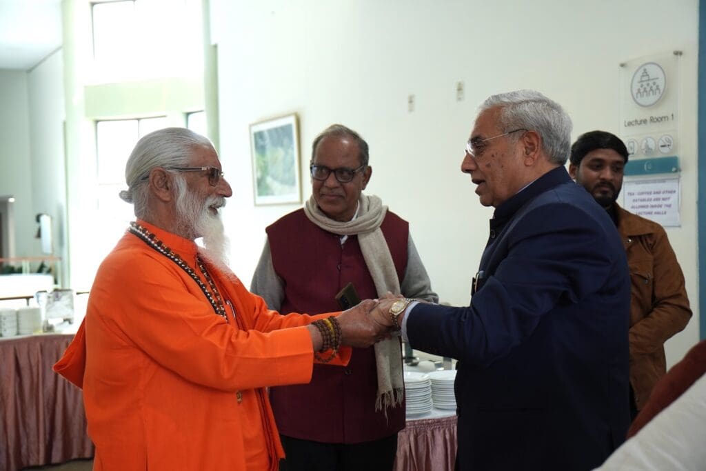 An elderly man in orange robes shakes hands with a man in a suit, symbolizing unity, while others look on in a room with tables and dishes in the background.