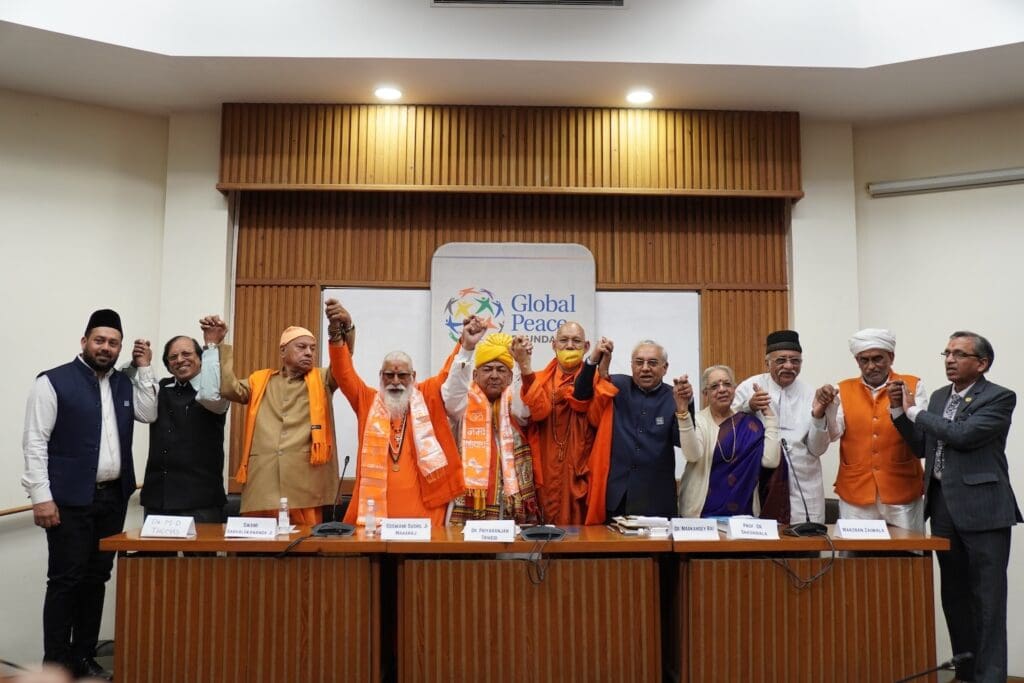 A group of religious and community leaders stand together at a table, holding hands above their heads in unity at an Interfaith Conclave during a Global Peace India event.