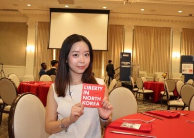 A person holds a "Liberty in North Korea" card while seated at a conference table set with red tablecloths in a banquet room.