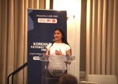Woman speaking at a podium with a microphone, in front of a banner that mentions "Korean Pathway to Peace" and sponsors.
