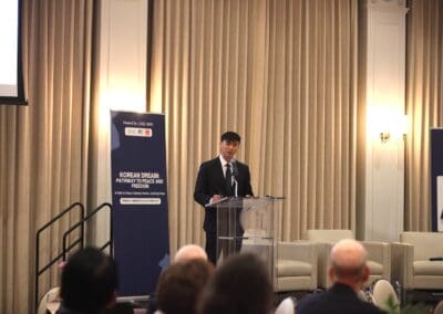 A speaker stands at a podium giving a presentation titled "Korean Dream: Pathway to Peace and Prosperity" in a conference room with an audience.