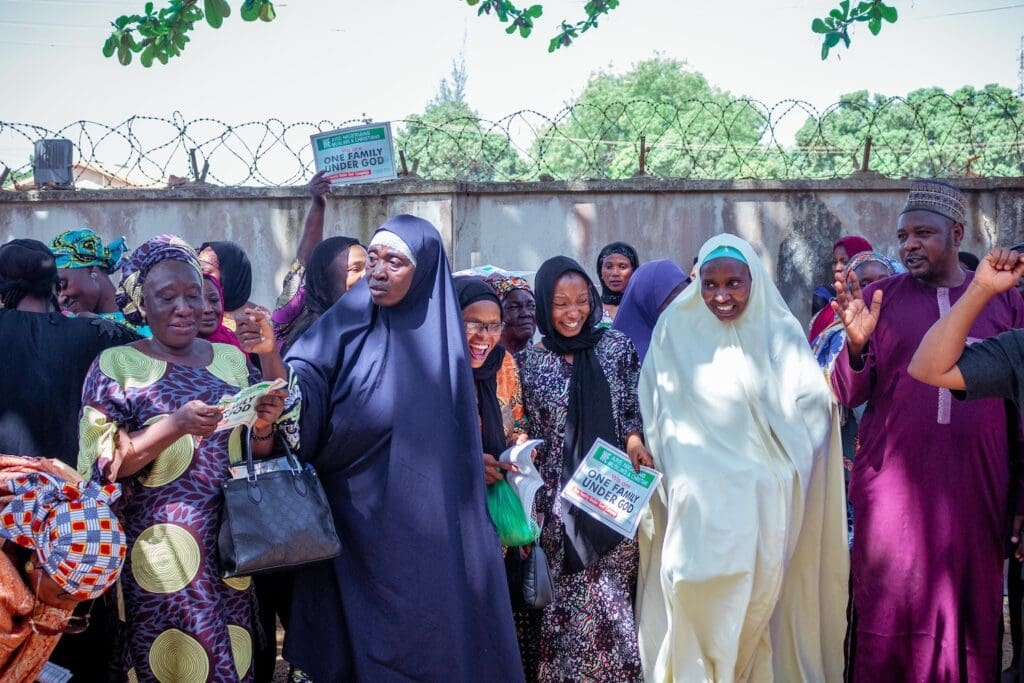A group of people, mostly women in colorful clothing and headscarves, stand outdoors near a fence, some holding signs and smiling while promoting Interfaith Harmony among Women in Nigeria.