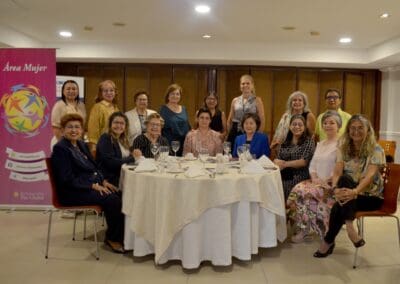 A group of women sit and stand around a round, set dining table in a banquet room; a banner labeled “Área Mujer Paraguay” is visible on the left, marking an International Women’s Day 2025 celebration.