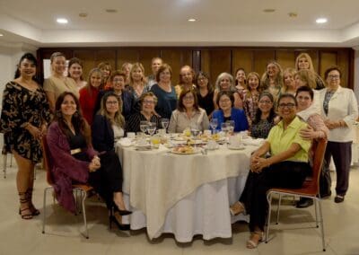A group of women pose for a photo around a round dining table set with glasses, plates, and food in a well-lit indoor setting, celebrating International Women’s Day with Area Mujer Paraguay.