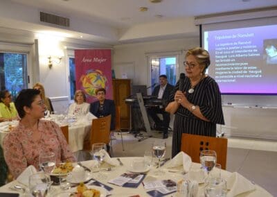 A woman speaks into a microphone at an International Women’s Day banquet event in 2025, with guests seated at tables, a musician playing keyboard, and a presentation by Area Mujer Paraguay projected on the wall behind her.