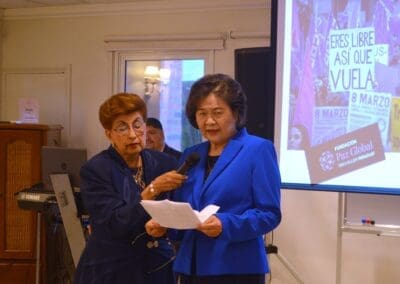 Two women in blue suits speak at an Area Mujer Paraguay event; one holds a microphone for the other, who reads from a paper. A presentation slide in Spanish is projected in the background, highlighting International Women’s Day.