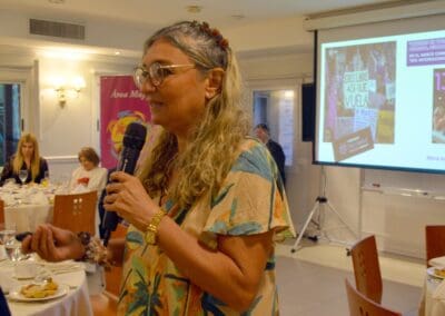 A woman with glasses speaks into a microphone at a formal indoor event for International Women’s Day, with a presentation projected on a screen behind her and people seated at tables.