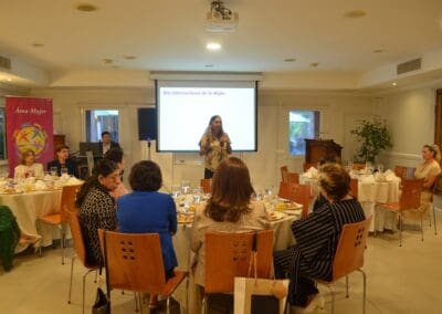 A woman stands and speaks into a microphone in front of a seated audience at a formal indoor event, with a presentation screen behind her during the International Women’s Day 2025 Celebration hosted by Area Mujer Paraguay.