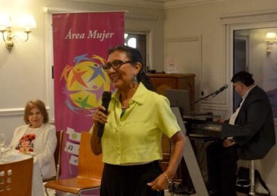 A woman in a yellow-green shirt speaks into a microphone at an indoor Area Mujer Paraguay event, with a musician at the keyboard. A pink "Área Mujer" banner decorates the 2025 Celebration for International Women’s Day.
