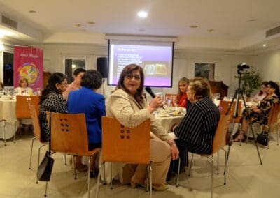 A woman holding a microphone speaks to a seated group during an indoor Area Mujer Paraguay meeting or event, with a presentation projected on a screen in the background, celebrating International Women’s Day 2025.