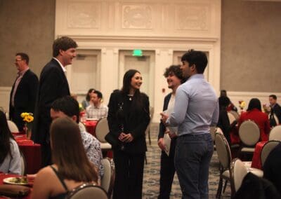 A group of people in formal attire stand and converse in a banquet hall with tables set for a meal.