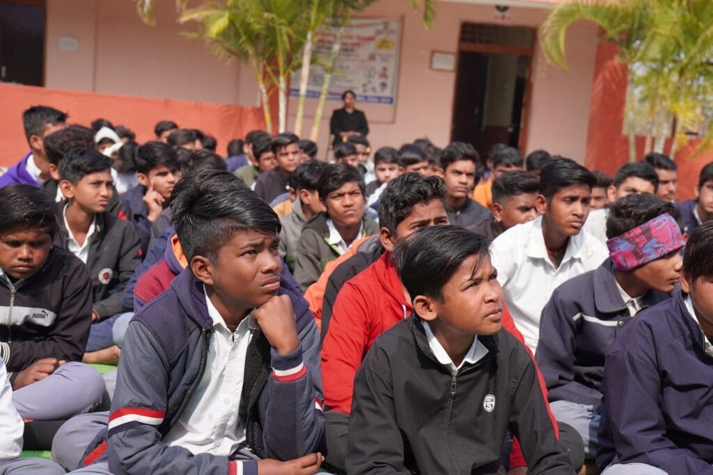 A group of students in uniforms sit outdoors on the ground, attentively listening as part of the PeaceHub Campaign, with a building and trees in the background, empowering youth for a better future.