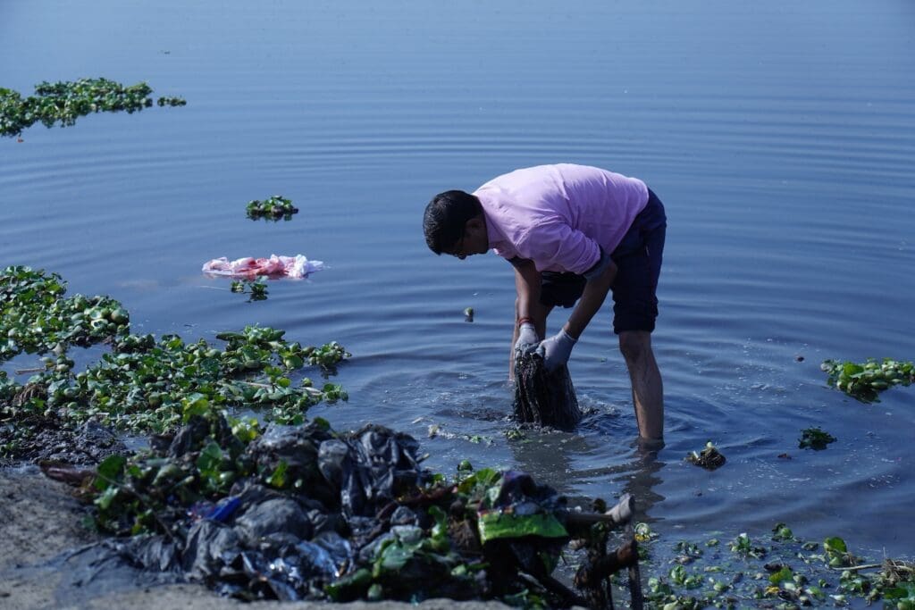 A person in a pink shirt diligently cleans trash from the Yamuna River, surrounded by floating plants and debris, as part of the 2025 GPF India clean-up initiative.