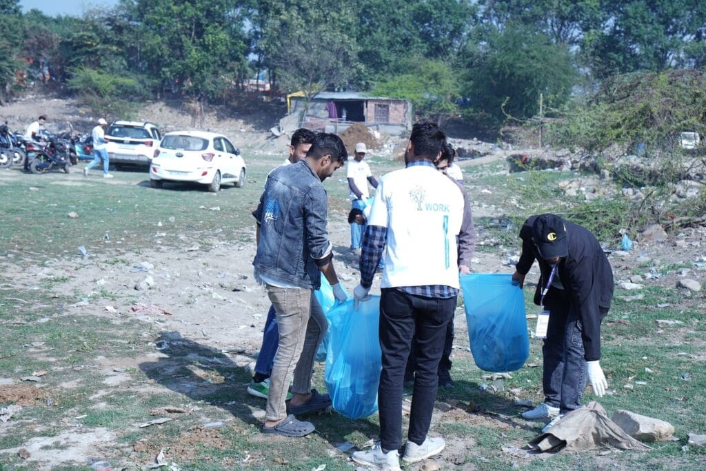 A dedicated group of people participated in a Clean-Up Drive, collecting trash in a grassy area with trees and parked vehicles in the background, as part of GPF India's efforts to preserve the Yamuna River's natural beauty.